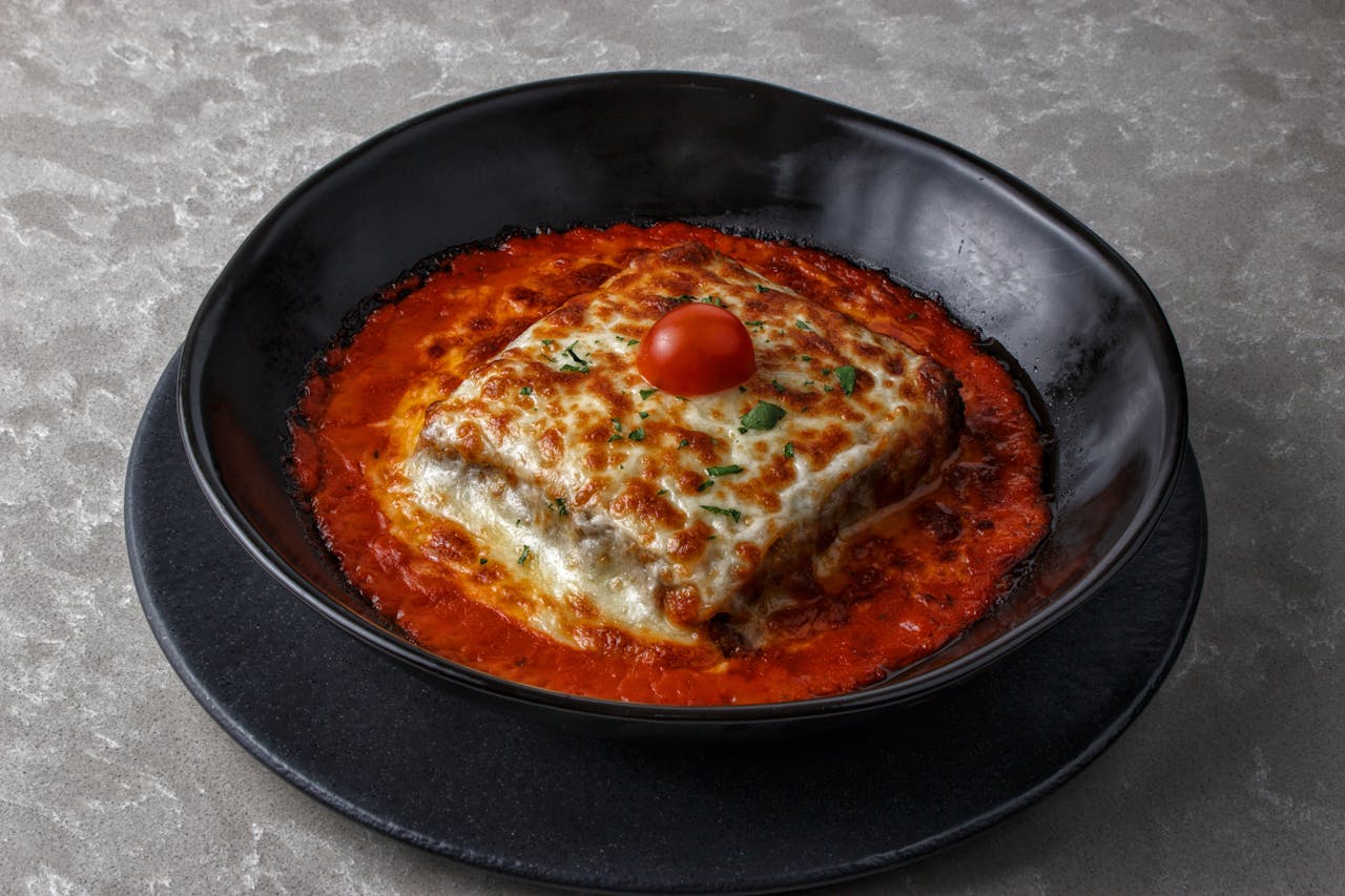 services-04 Tasty baked lasagna with cherry tomato and cheese, served in a black bowl with rich tomato sauce.