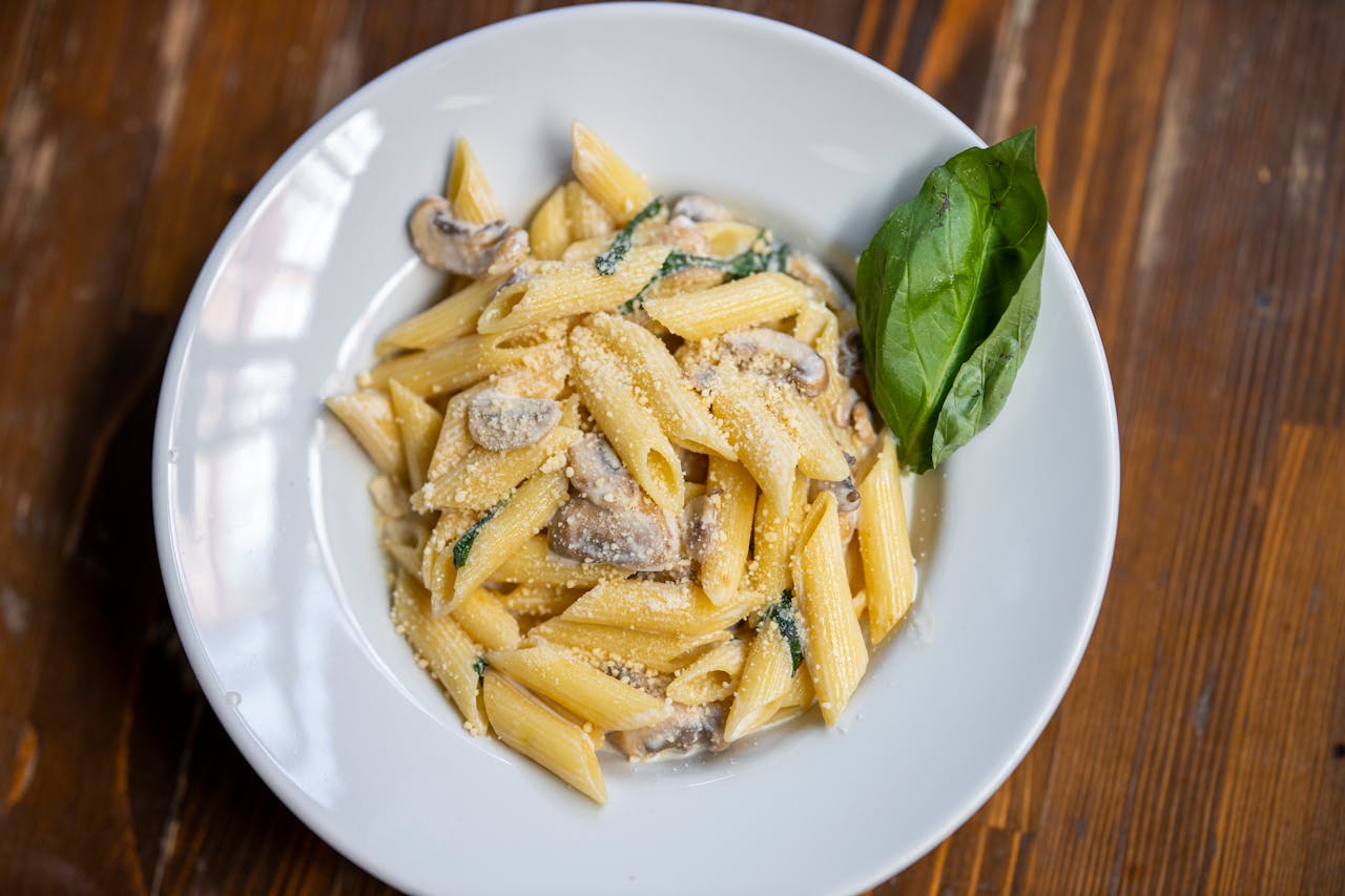 contact-img Delicious creamy penne pasta garnished with basil, mushrooms, and parmesan cheese on a rustic wooden table.